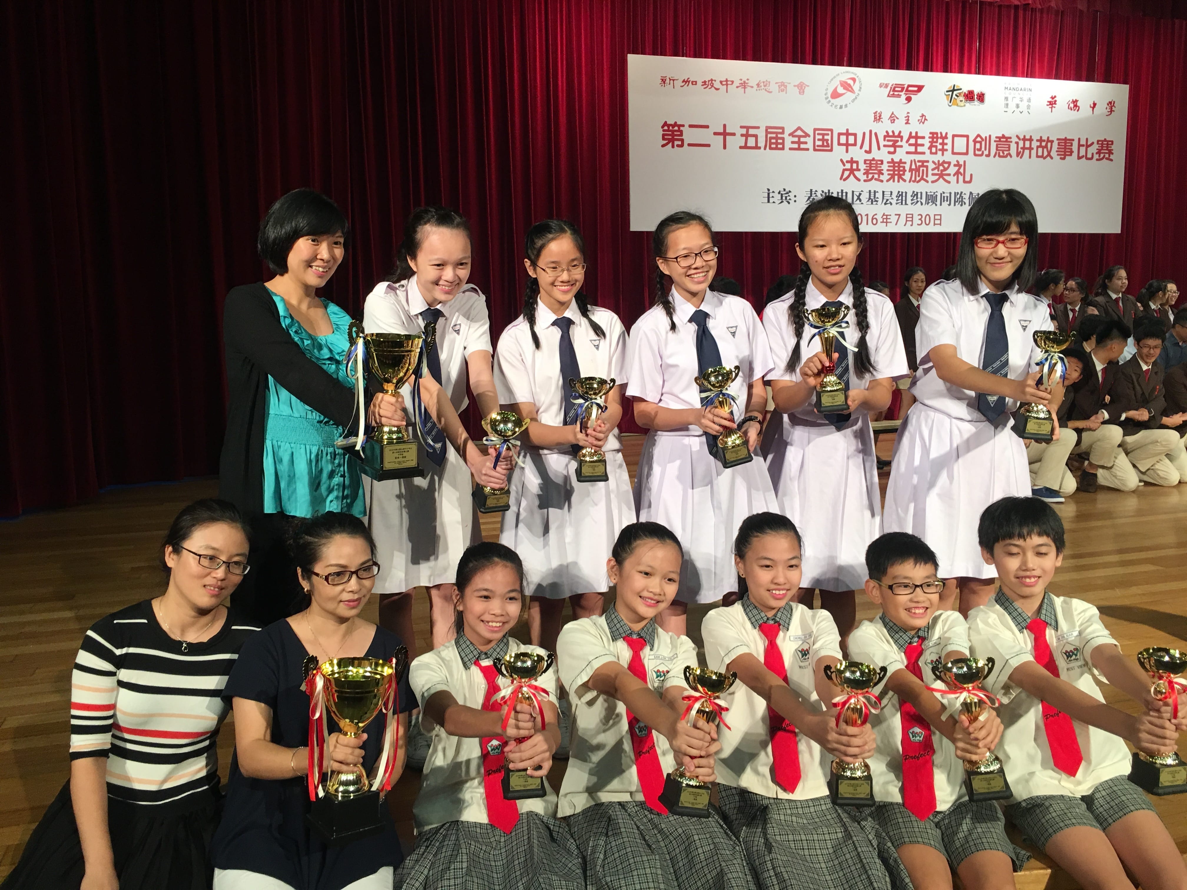 Students in school uniforms pose with gold trophies in front of a red curtain and a Chinese competition banner.